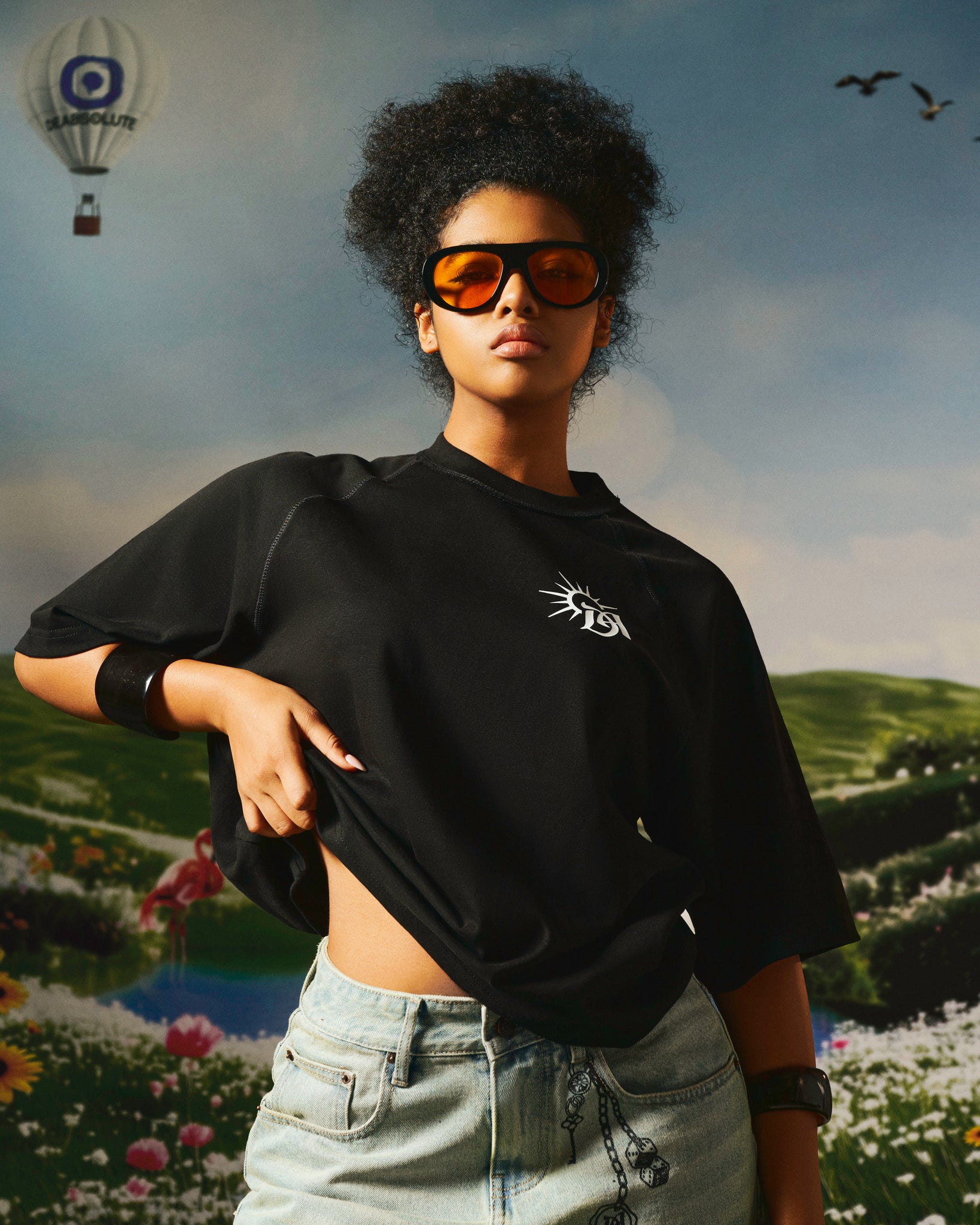 Person wearing a black crop top with a logo, standing against a scenic background with balloons and birds.